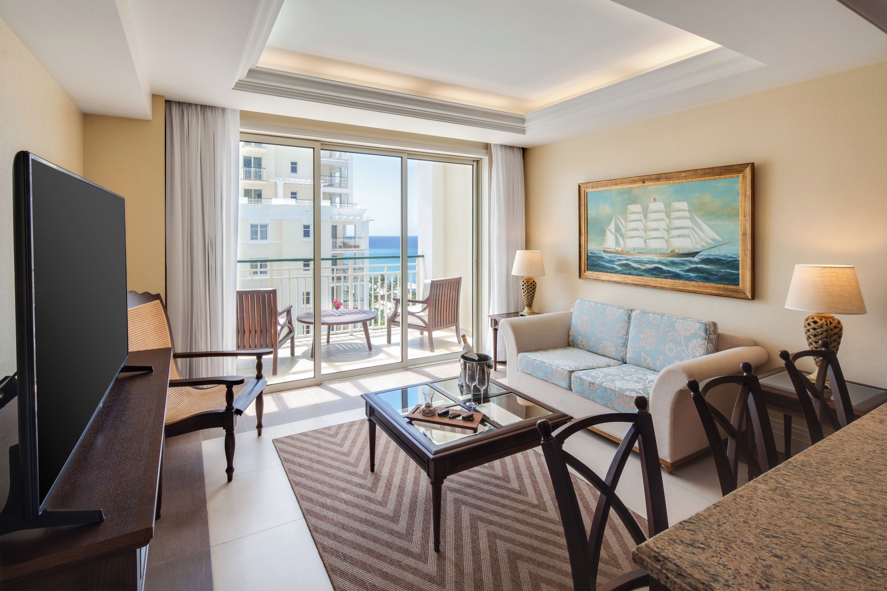 Habitación de hotel con balcón, televisión, sofá, mesa de centro y un cuadro de un barco.