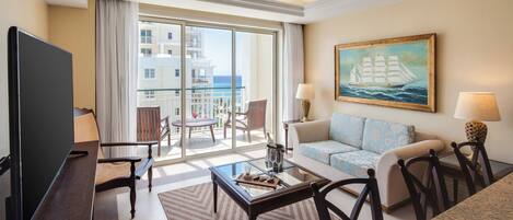 Habitación de hotel con balcón, televisión, sofá, mesa de centro y un cuadro de un barco.
