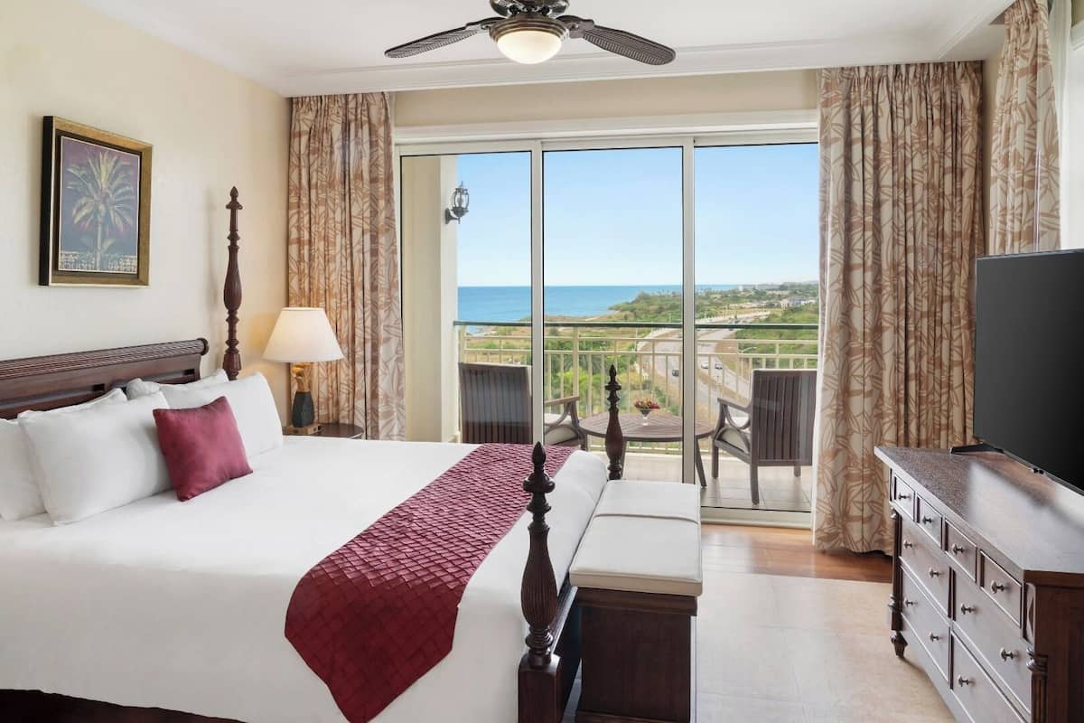 Una habitación de hotel con cama, televisión, balcón y vista al océano, además de ventilador de techo.