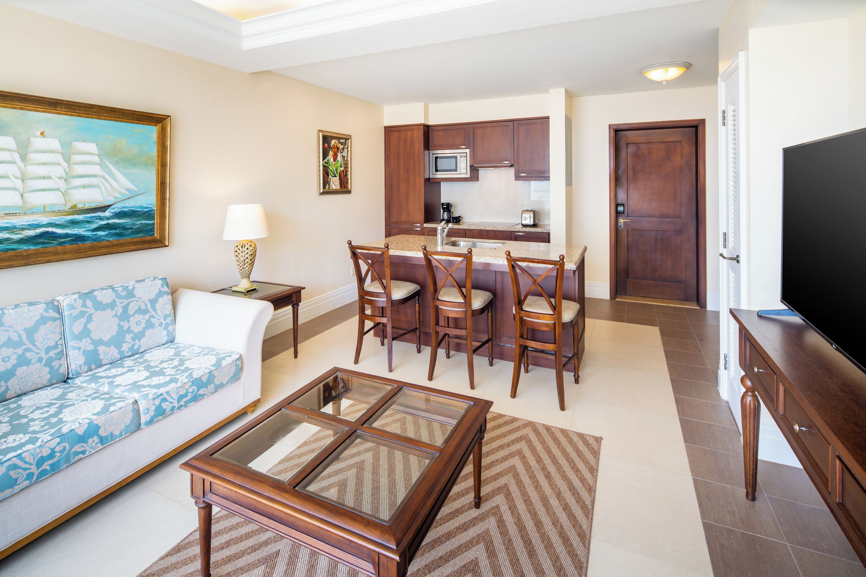 A living room with a sofa, a coffee table, and a television. There is a kitchen area with a microwave, a coffee maker, and a toaster. The room has wooden cabinets and a door.