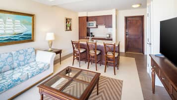 A living room with a sofa, a coffee table, and a television. There is a kitchen area with a microwave, a coffee maker, and a toaster. The room has wooden cabinets and a door.