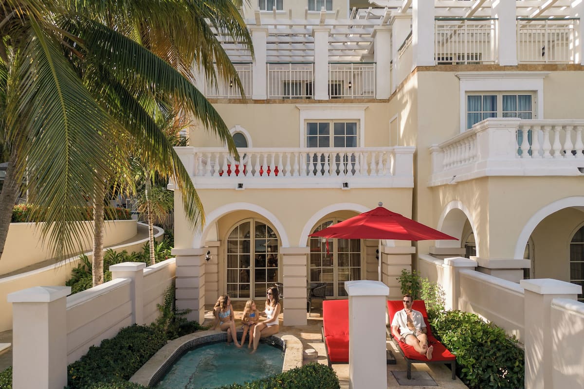 Zona de piscina con sillas de descanso rojas, una sombrilla roja y una piscina rodeada de follaje y un edificio blanco con balcones.