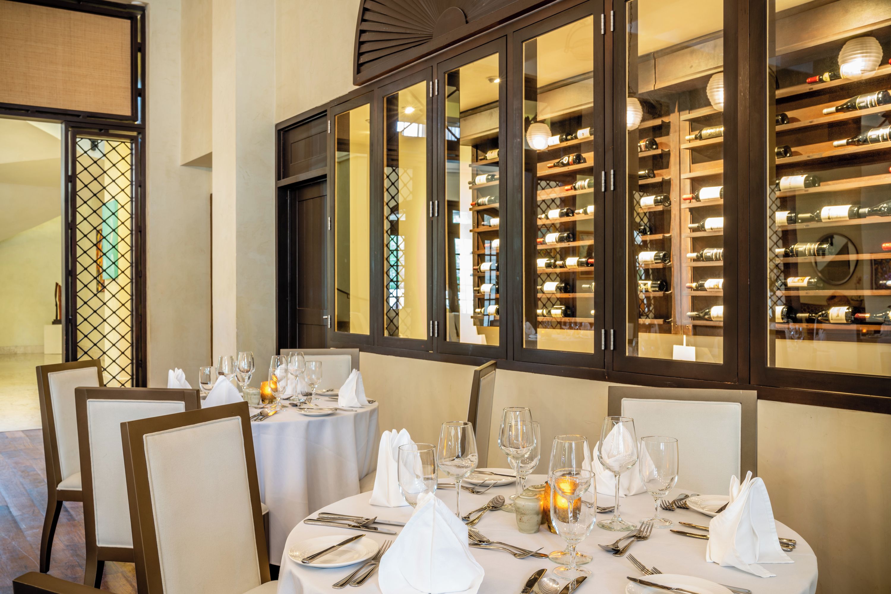 A dining area with tables set for a meal, chairs, and a wine rack visible through a window.