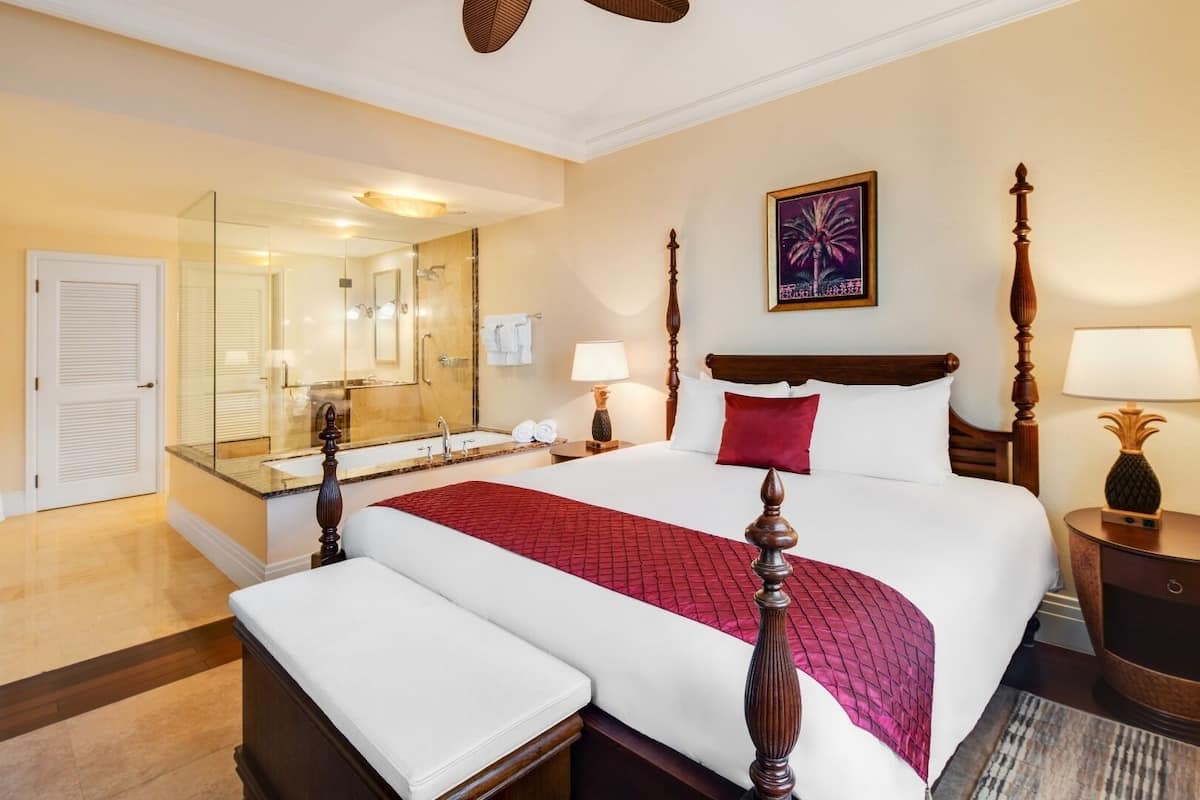 Habitación de hotel con una cama grande, un área de estar separada, un baño con cabina de ducha de vidrio y un cuadro en la pared.