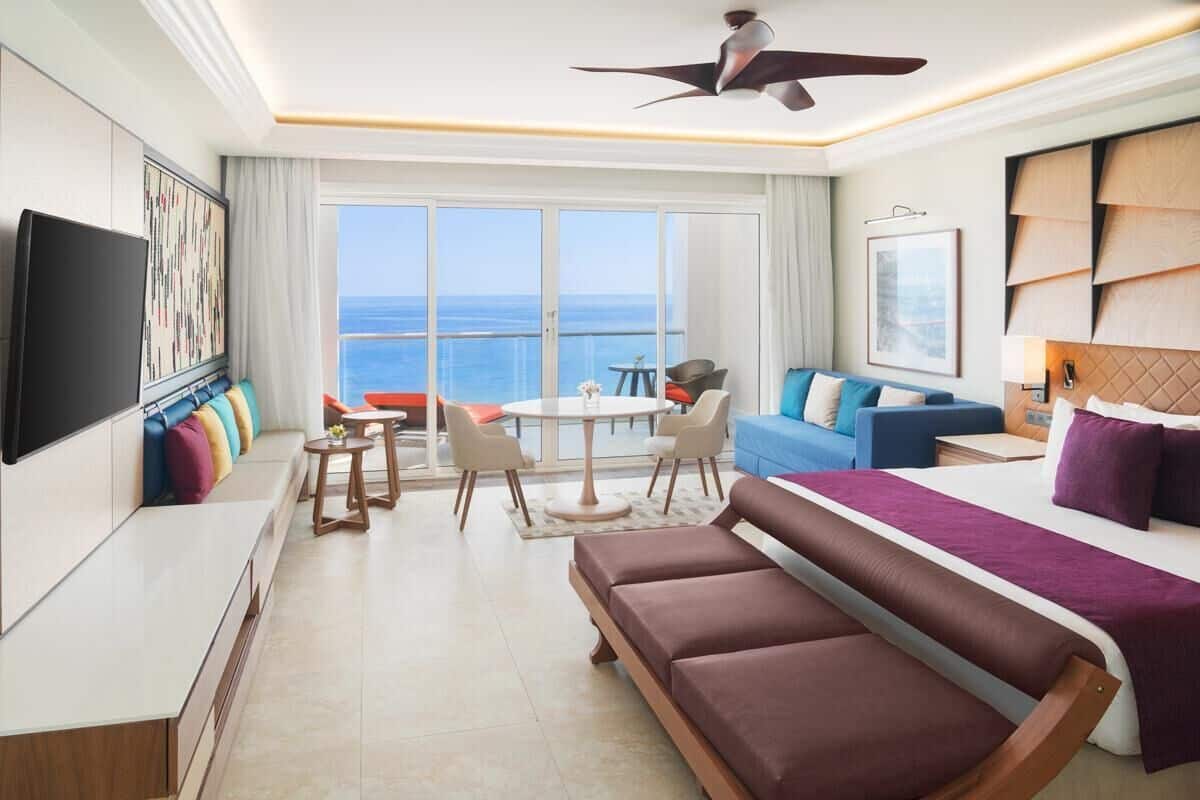 Una habitación de hotel moderna con una cama grande, un sofá, un televisor, un balcón con vistas al mar y un ventilador de techo.