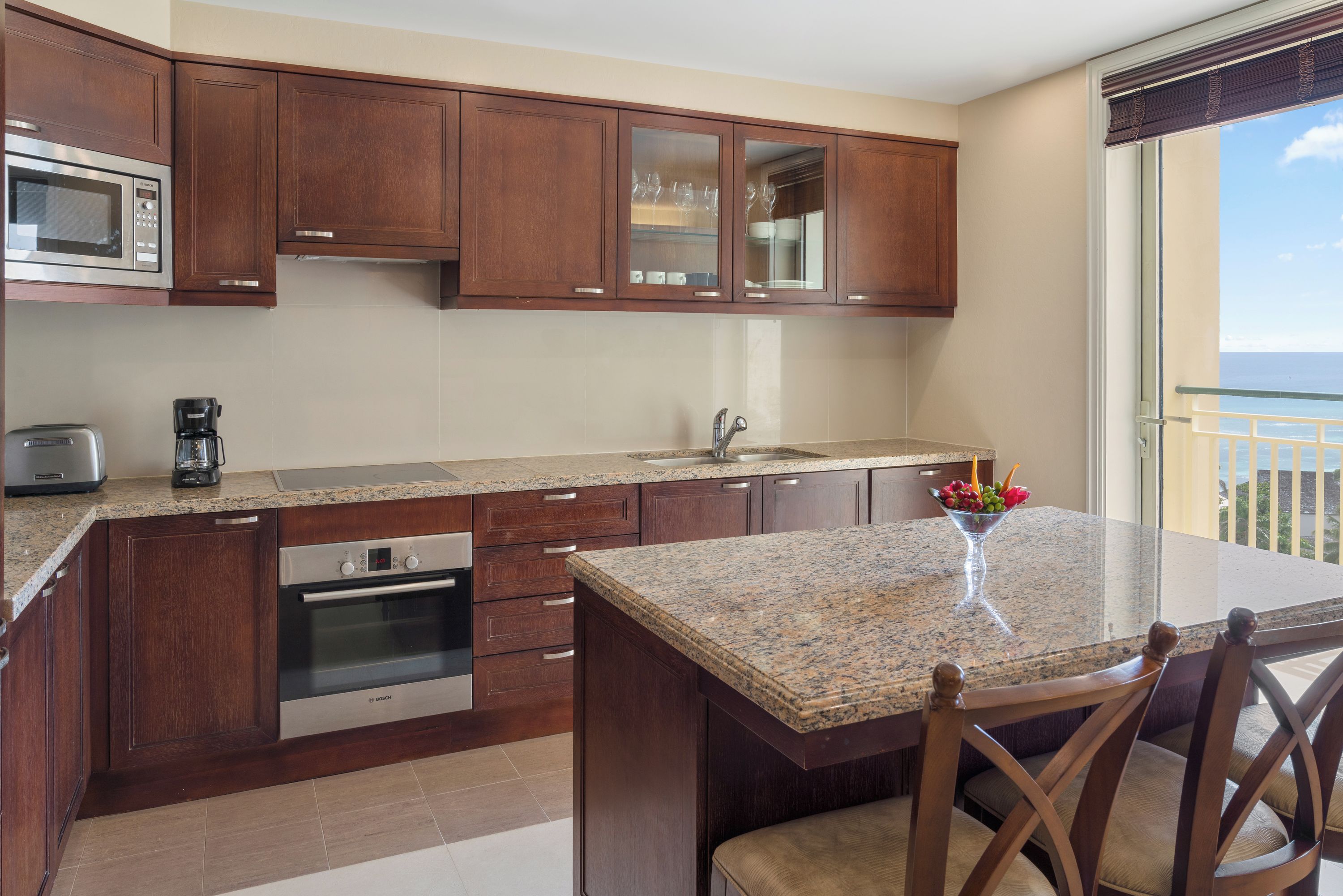A modern kitchen with wooden cabinets, a granite countertop, a built-in oven, a coffee maker, and a view of the ocean through a balcony.