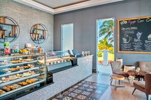 Una cafetería con una vitrina de pasteles, un menú en pizarra y un área de descanso con vista a palmeras.