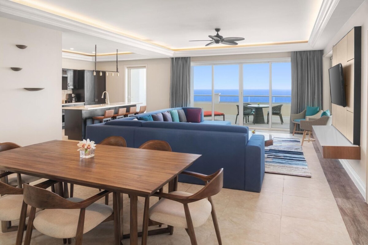 A modern living room with a large blue sofa, a wooden dining table with chairs, a kitchen area, and a balcony with a view of the ocean.