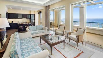 A spacious living room with a sofa, armchairs, and a coffee table. There is a kitchen area in the background and large windows with a view of the ocean.
