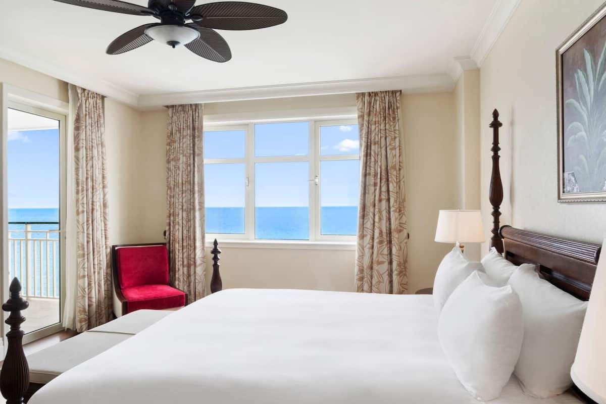 Una habitación de hotel con una cama grande, una silla roja, un ventilador de techo y vista al mar a través de una ventana.