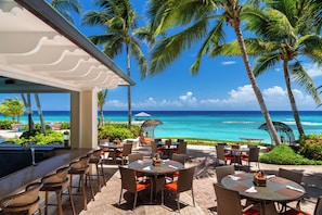 An outdoor dining area with tables and chairs, overlooking a beach with palm trees and the ocean.
