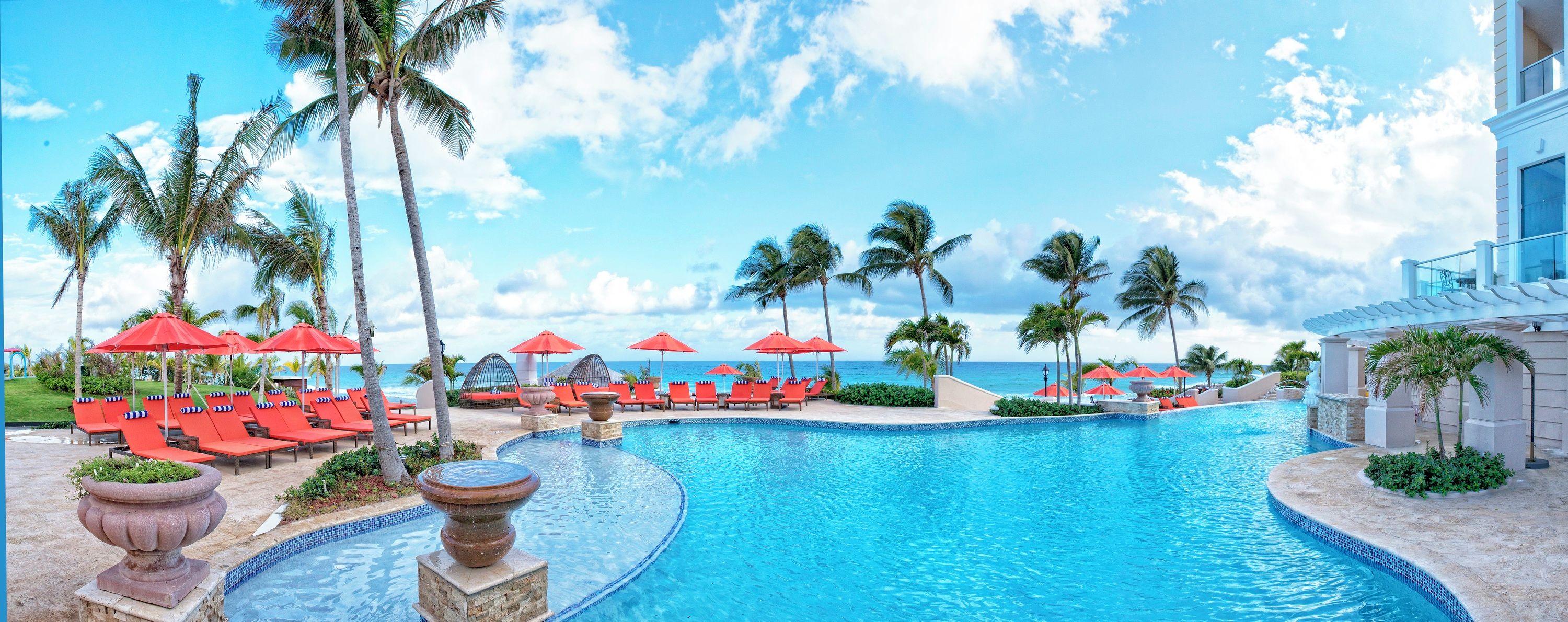 A pool with red umbrellas and lounge chairs, surrounded by palm trees and a clear sky.