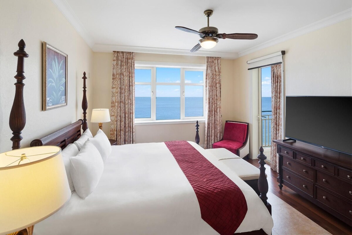 A hotel room with a large bed, a television, a ceiling fan, and a view of the sea.