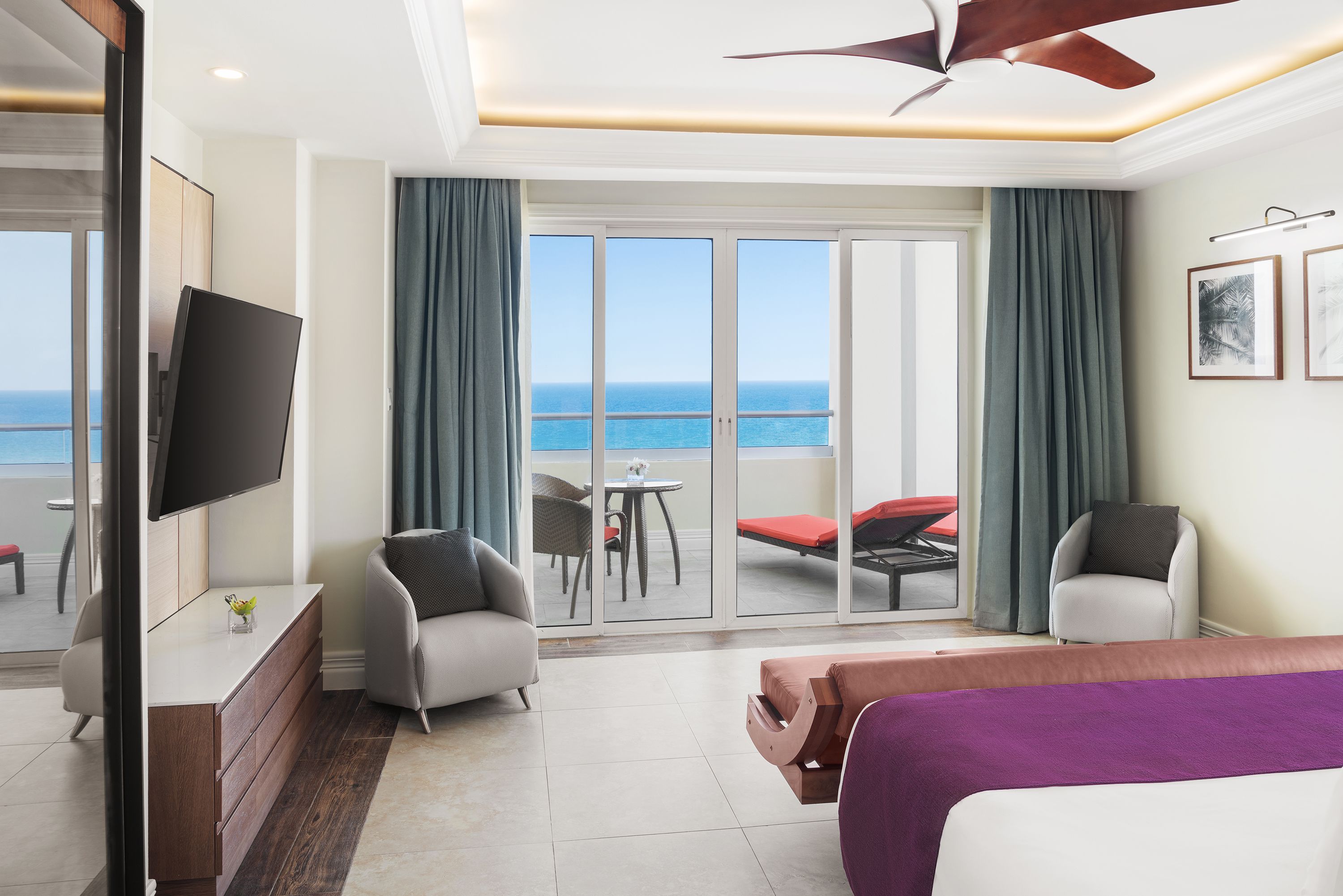 A hotel room with a bed, a desk, a TV, a balcony with a view of the sea, and two chairs.