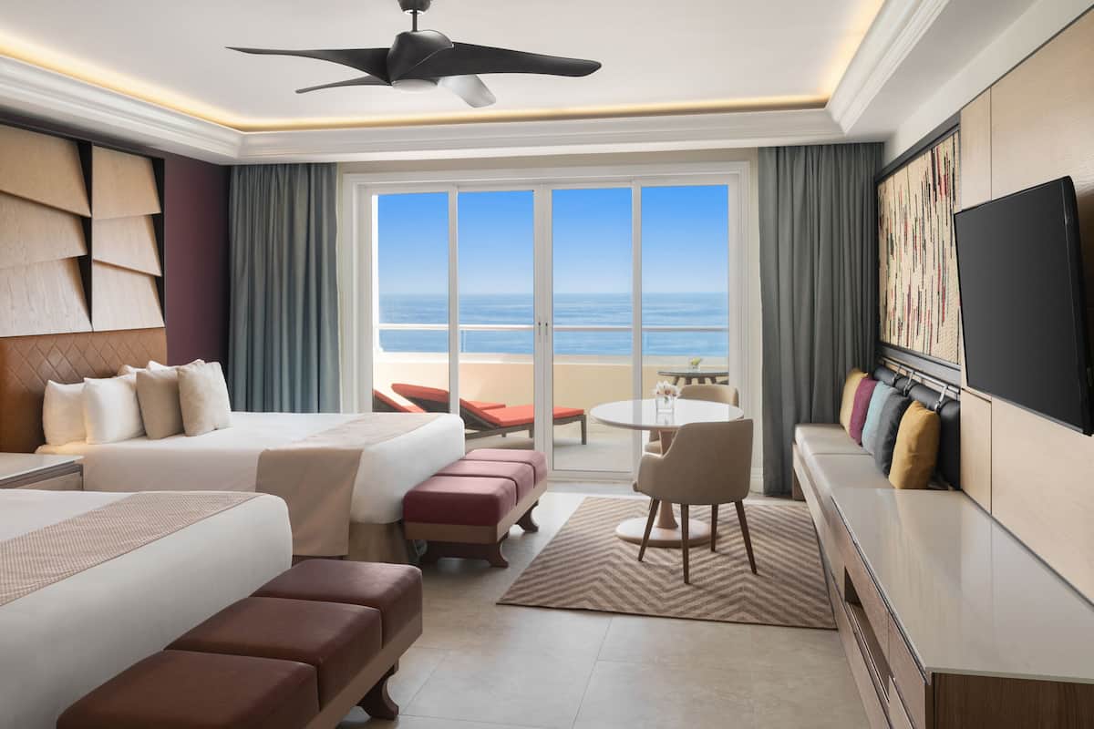 Habitación de hotel con cama, zona de estar con mesa y sillas, televisor y vista al océano.