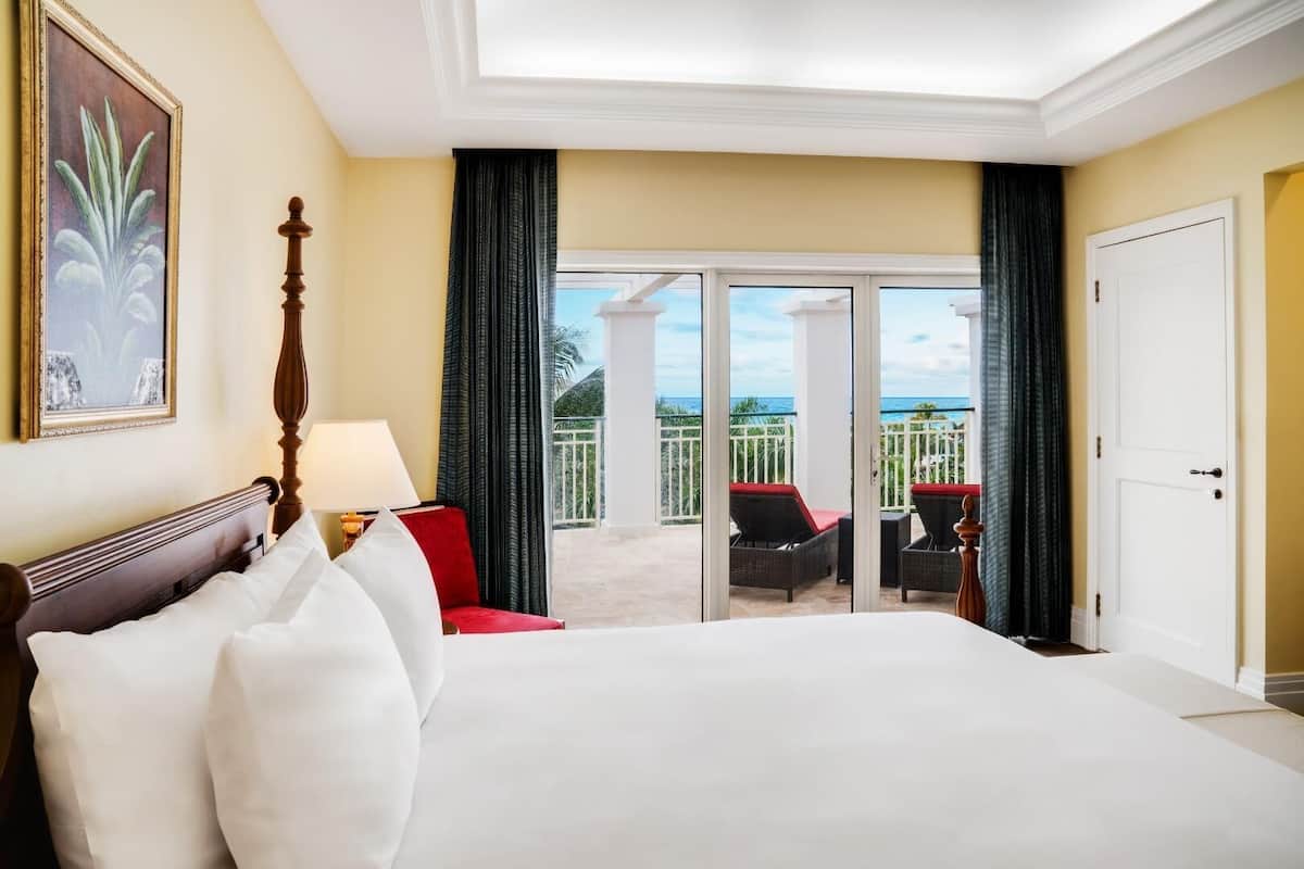 Habitación de hotel con una cama grande, una silla roja, un balcón con mobiliario de mimbre y vistas al océano.