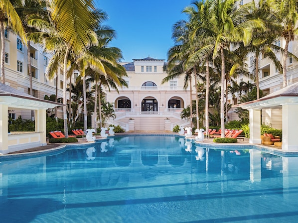 A swimming pool with lounge chairs, palm trees, and a building with arched windows and a balcony.