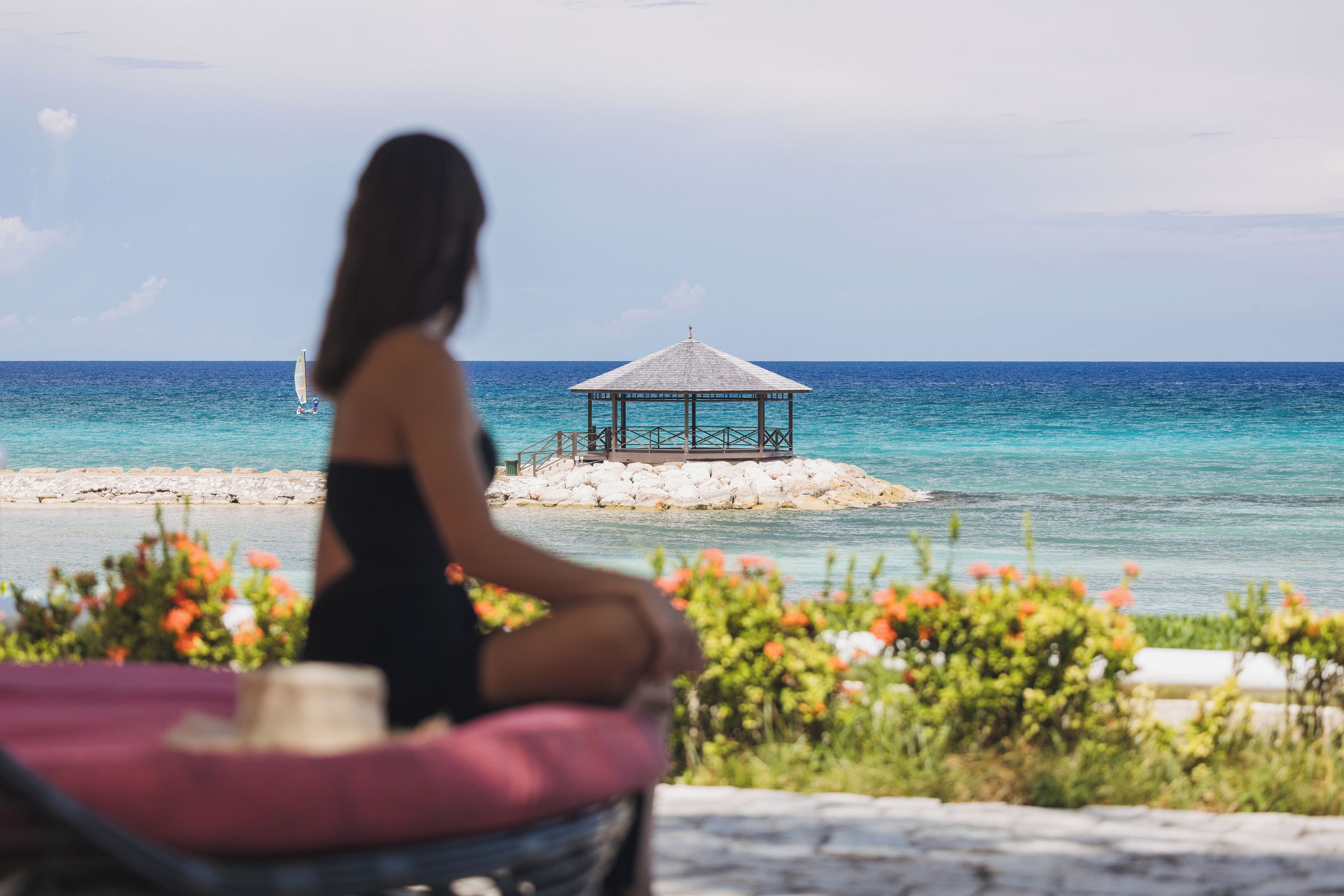 A person sitting on a chair with a view of the ocean and a gazebo on a rocky outcrop.