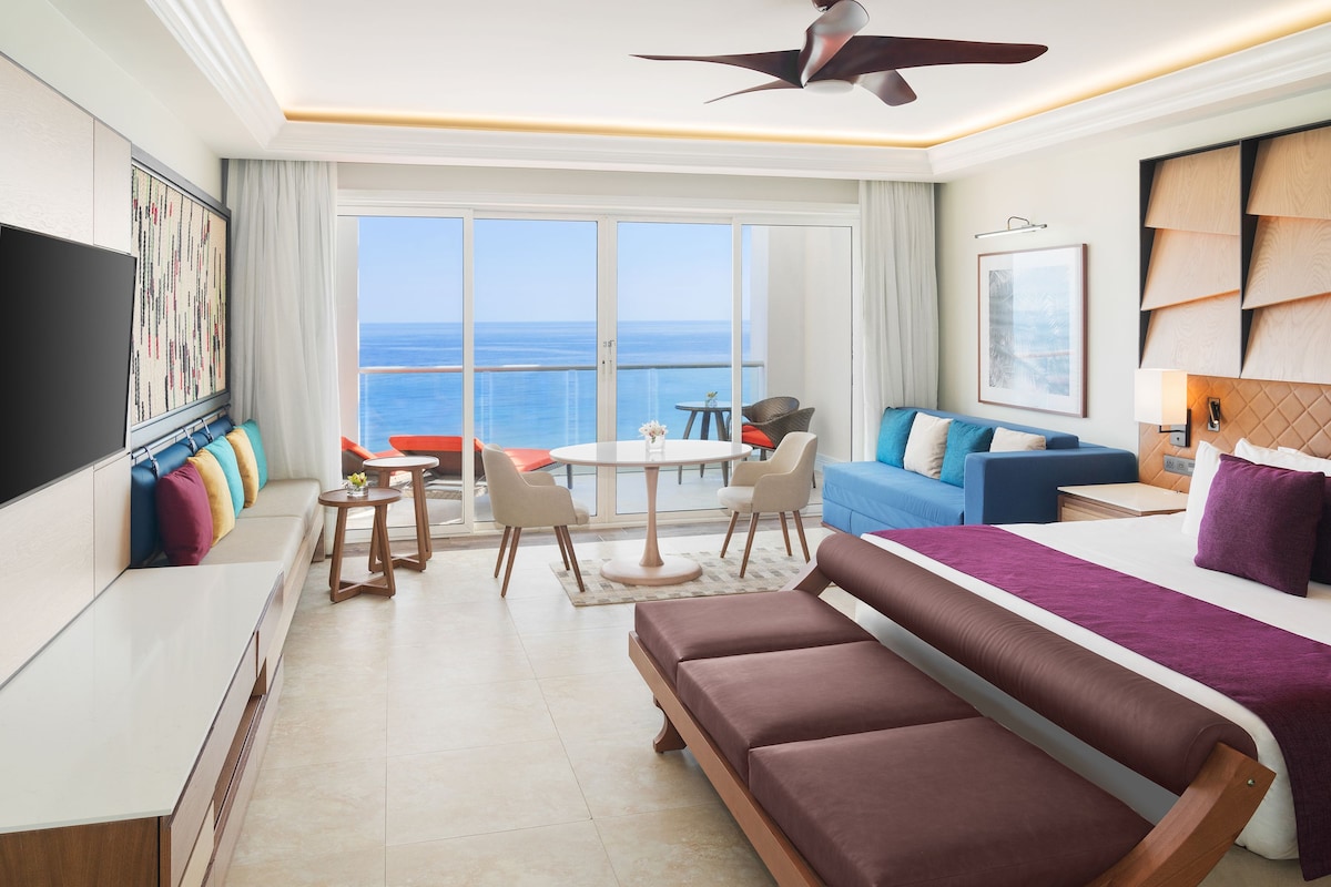 A modern hotel room with a large balcony, a sofa, a bed, a TV, and a dining area.