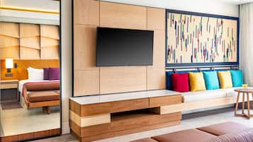 A modern hotel room with a large mirror, a flat-screen TV mounted on a wooden panel, a sofa with colorful cushions, and a large abstract artwork on the wall.