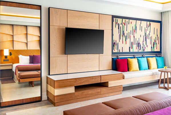 Une chambre d’hôtel moderne dotée d’un grand miroir, d’un téléviseur à écran plat fixé sur un panneau en bois, d’un canapé agrémenté de coussins colorés et d’une grande œuvre d’art abstraite accrochée au mur.