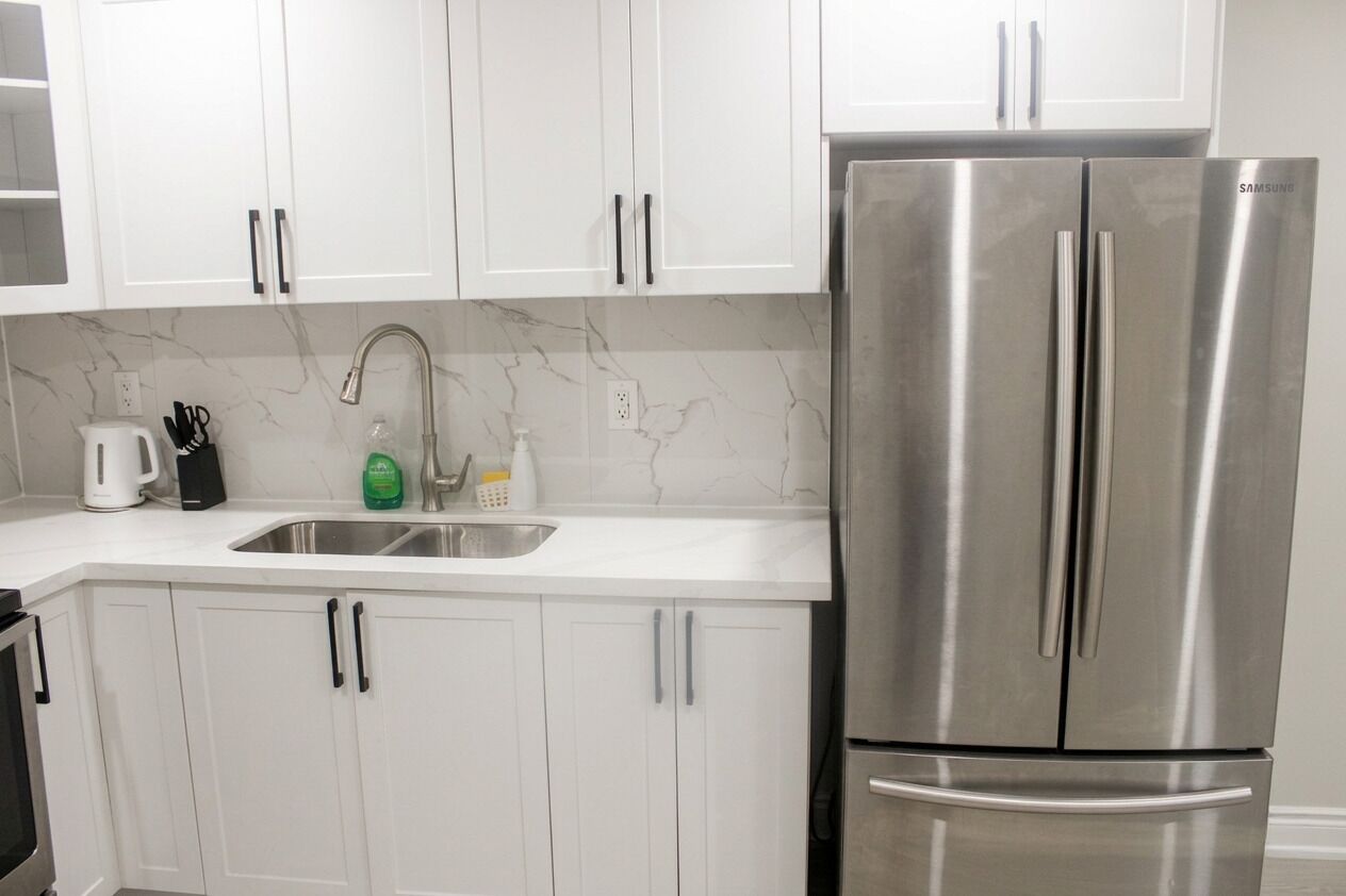 Una cocina con armarios blancos, un refrigerador de acero inoxidable, un lavaplatos y un protector contra salpicaduras de mármol.