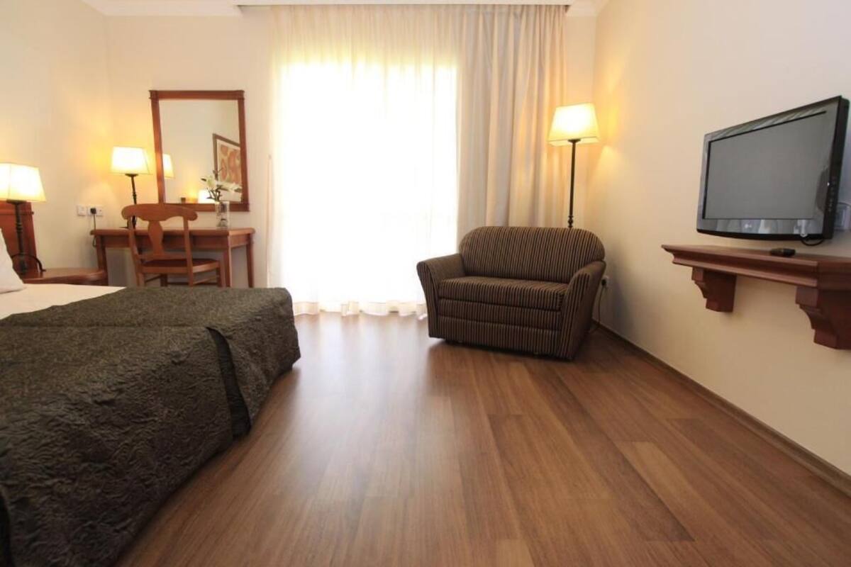 Habitación de hotel con cama, una silla, escritorio con lámpara, televisor y una ventana grande con cortinas.