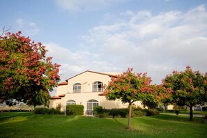 A two-story building with a flat roof, surrounded by well-maintained grass and trees with pink flowers. - Pastoral Hotel (HaGalil HaElyon)