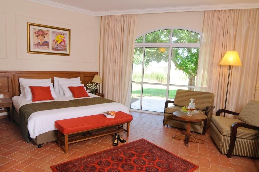 Quarto de hotel com cama grande, um banco, uma cadeira, uma mesa e janela com cortinas.