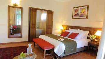Una habitación de hotel con cama, lámparas de noche, un banco, una mesita con una bandeja de frutas y una botella, y un espejo.