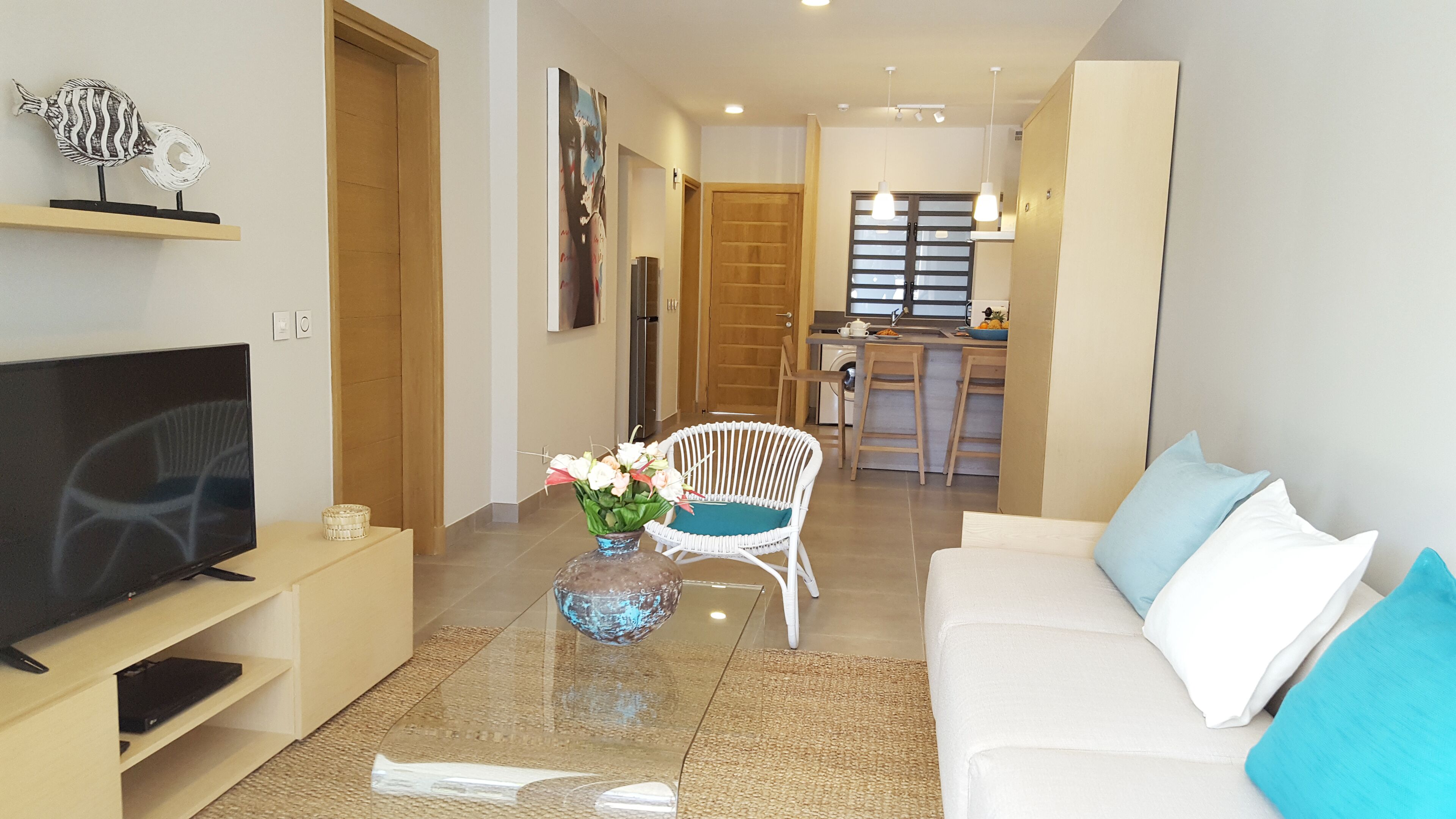 A modern living room with a TV, a glass coffee table, a white sofa, and a wicker chair. There is a kitchen area in the background with a dining table and chairs.