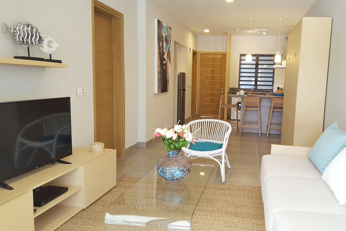 Una sala moderna con televisor, una mesa de centro de vidrio, un sofá blanco y una silla de mimbre. En el fondo, se distingue una zona de cocina con una mesa y sillas de comedor.