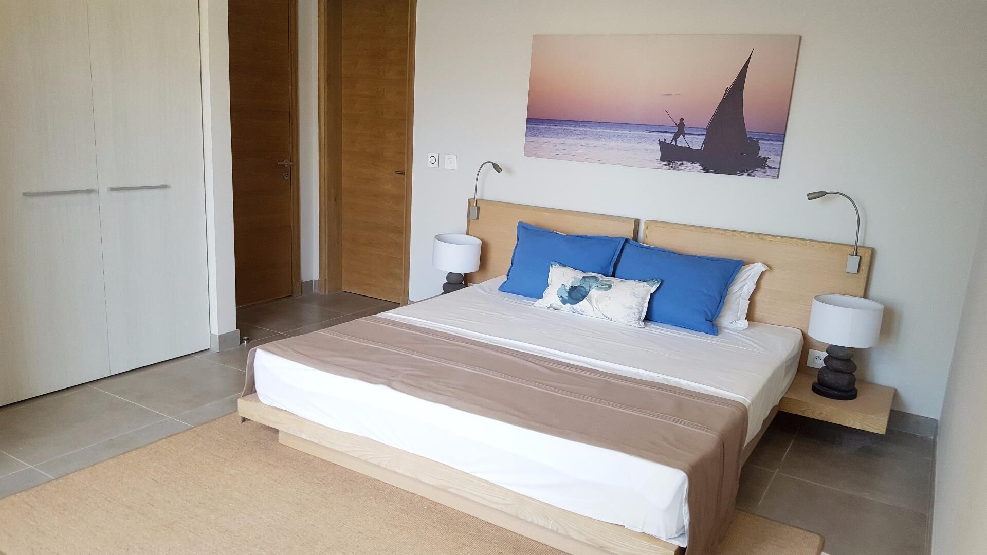 A bedroom with a bed, two bedside lamps, a painting of a sailboat, and a wooden door.