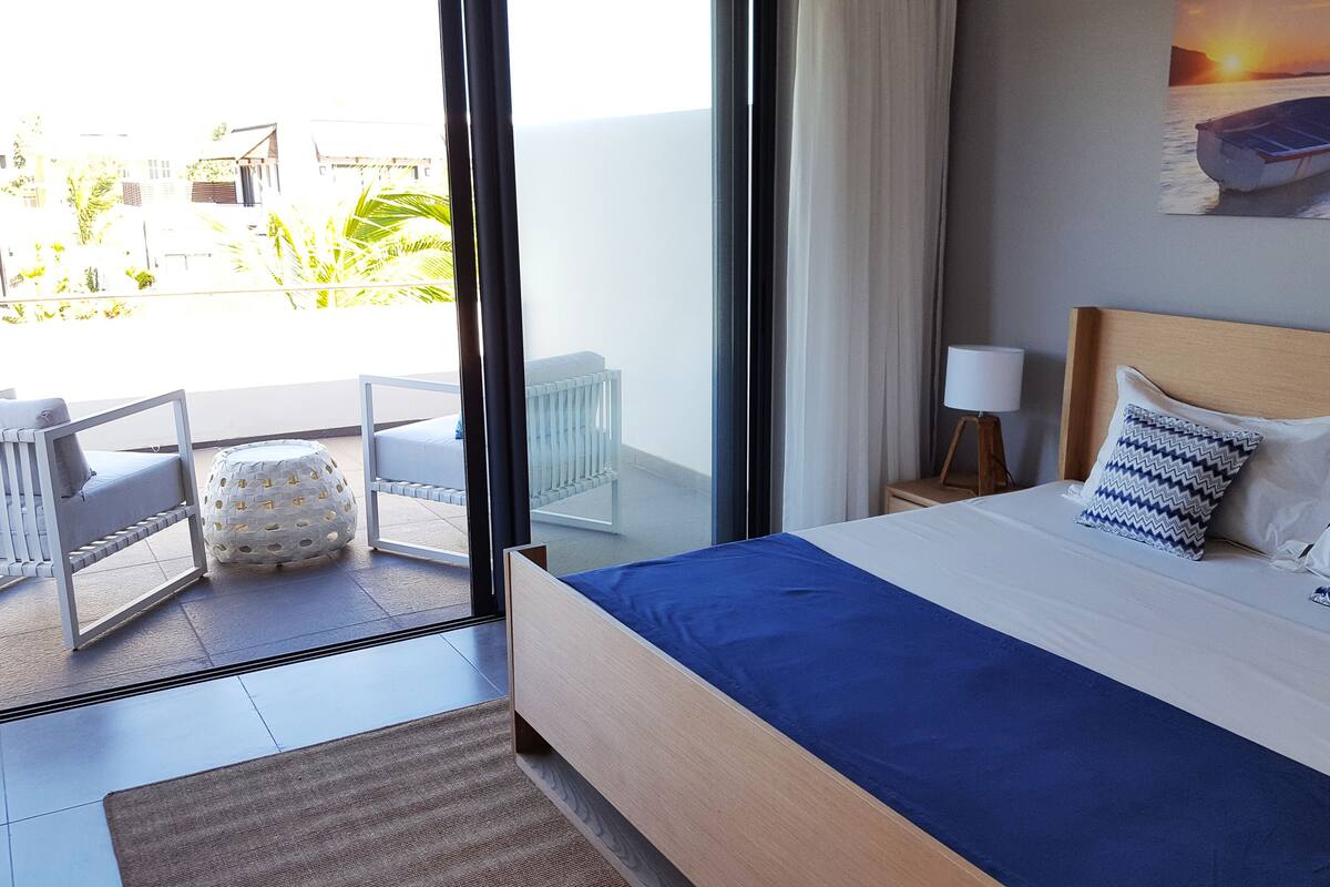 Habitación de hotel con cama, mesita de noche, lámpara, vista al mar y balcón con mobiliario exterior.