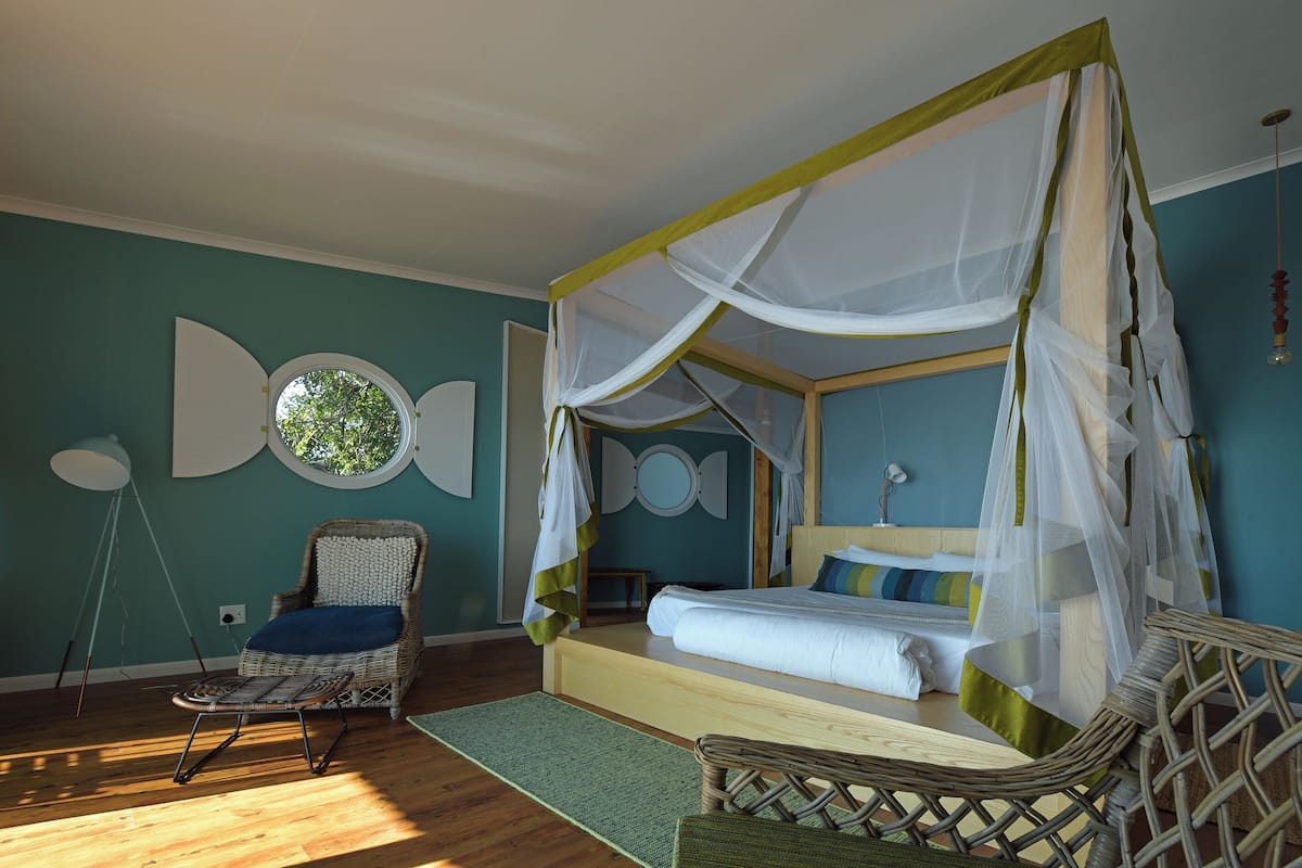 Un dormitorio con cama con dosel, una silla de mimbre, una mesita y un espejo redondo en la pared.