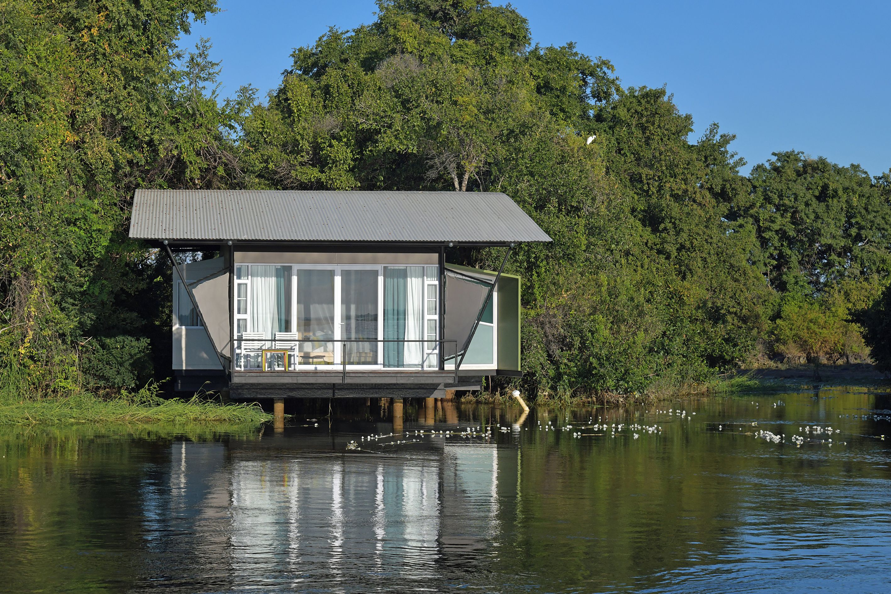 Een drijvend huis met een groot raam, gelegen op het water, omgeven door bomen.