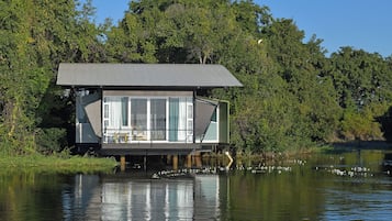 Een drijvend huis met een groot raam, gelegen op het water, omgeven door bomen.