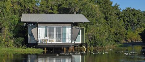 A floating house with a large glass window, situated on a body of water surrounded by trees.