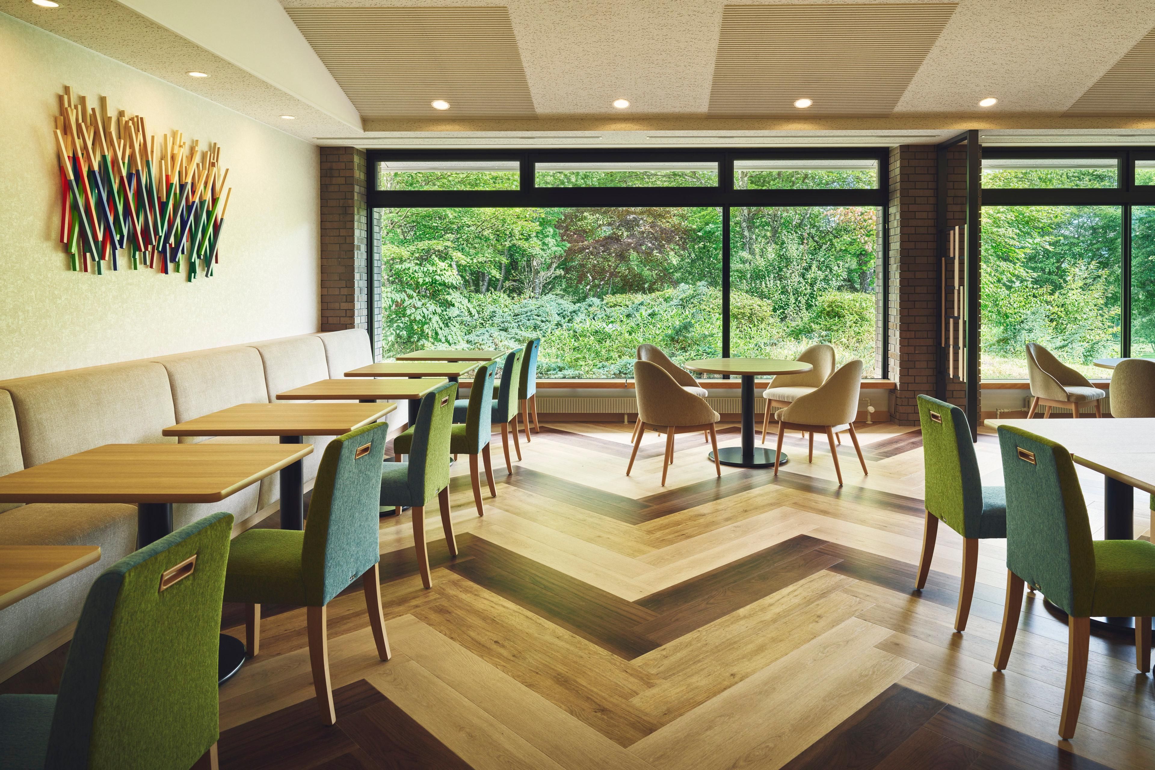A modern dining area with wooden tables and chairs, large windows with a view of greenery, and a unique wall art installation.