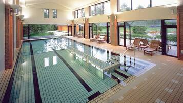 A swimming pool with a glass barrier, wooden deck chairs, and a view of greenery outside.