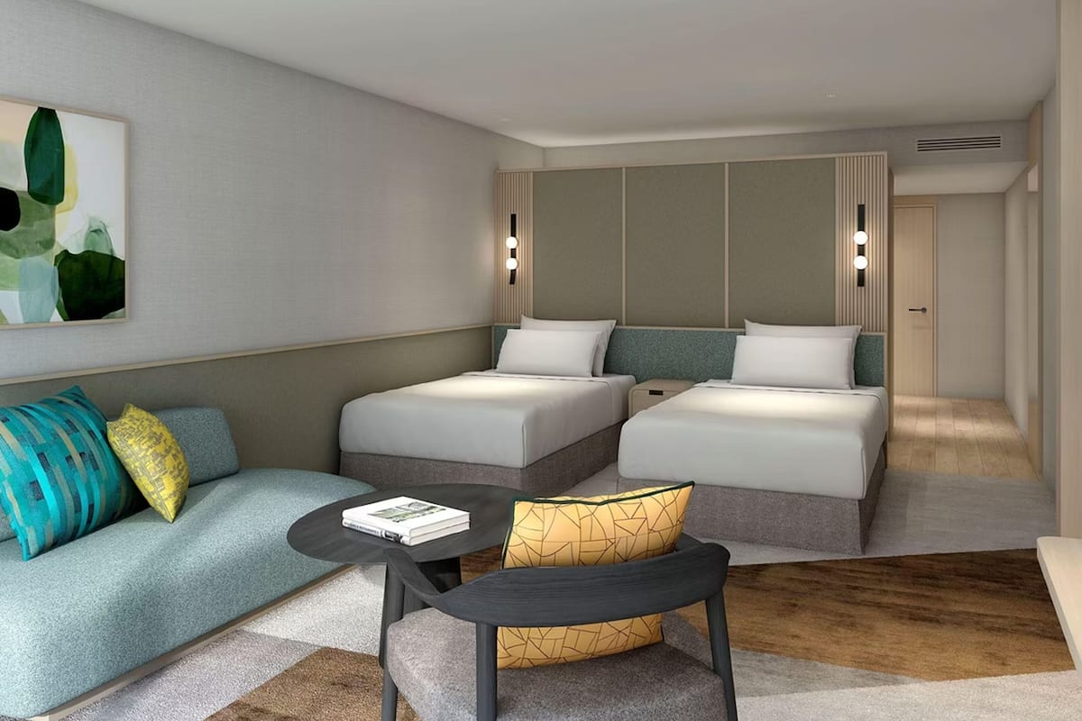 Moderni hotellihuone, jossa on sohva, kaksi sänkyä, pieni pöytä ja seinään kiinnitetty valaisin.