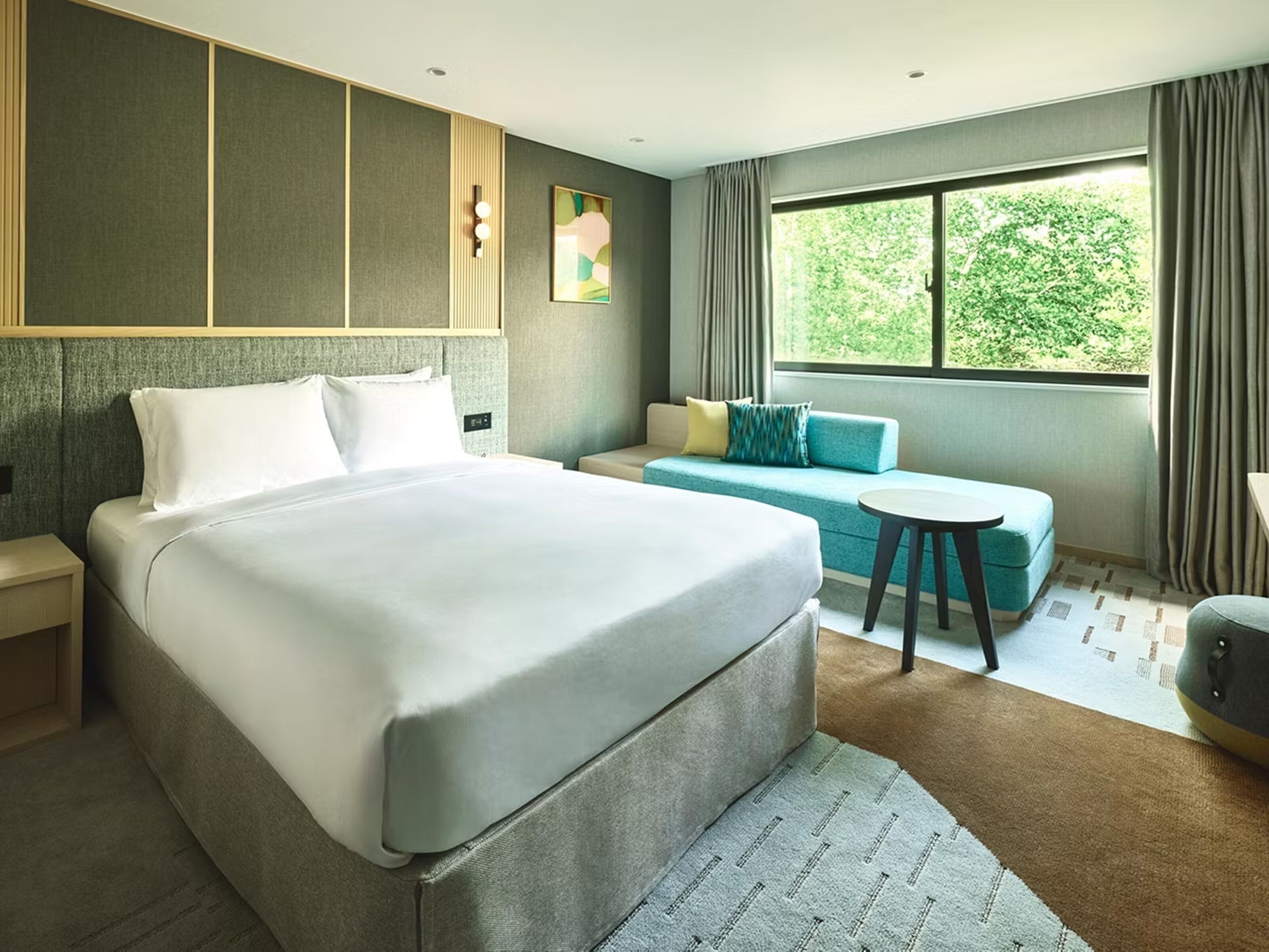 Ein modernes Hotelzimmer mit einem großen Bett, einer Couch, einem kleinen runden Tisch und einem Fenster mit Blick auf Grünflächen.