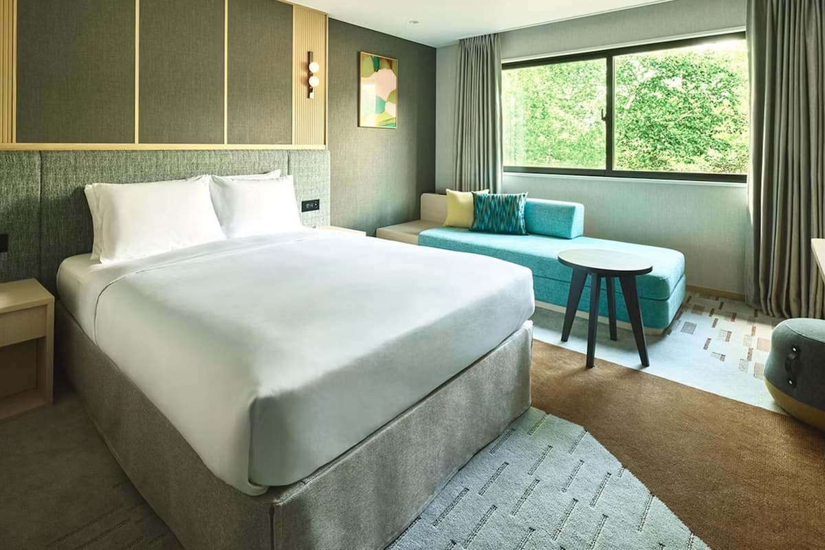 Moderni hotellihuone, jossa on suuri sänky, sohva, pieni pyöreä pöytä ja ikkuna, josta avautuu näkymä vehreään maisemaan.