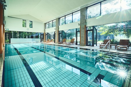 A large indoor swimming pool with clear glass walls, wooden lounge chairs, and a view of greenery outside.