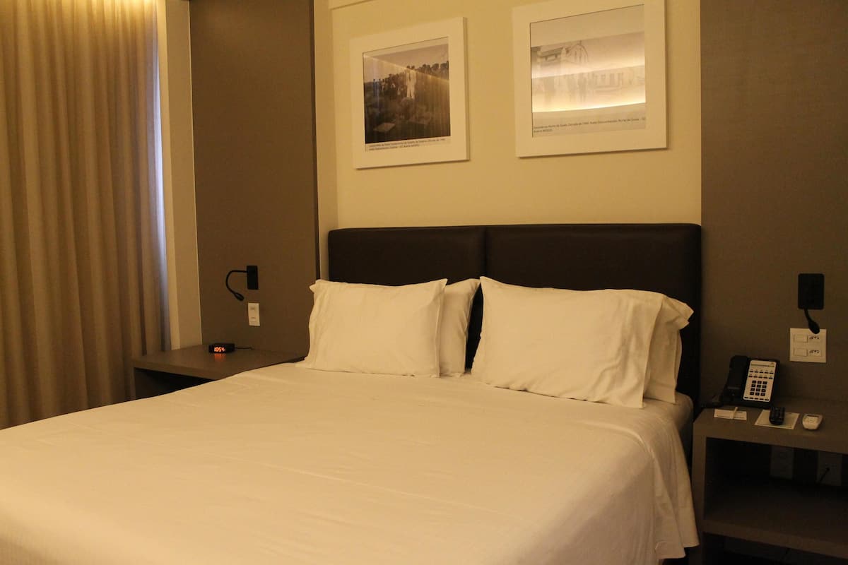 Habitación de hotel con una cama grande, dos almohadas, una mesita de noche con teléfono y lámpara, y dos cuadros en la pared.