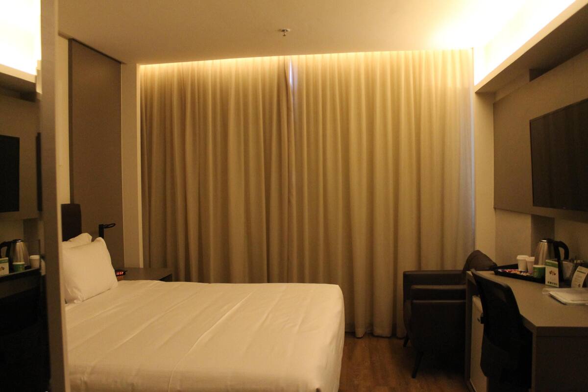 Una habitación de hotel con cama, escritorio, silla, televisor y cortina.