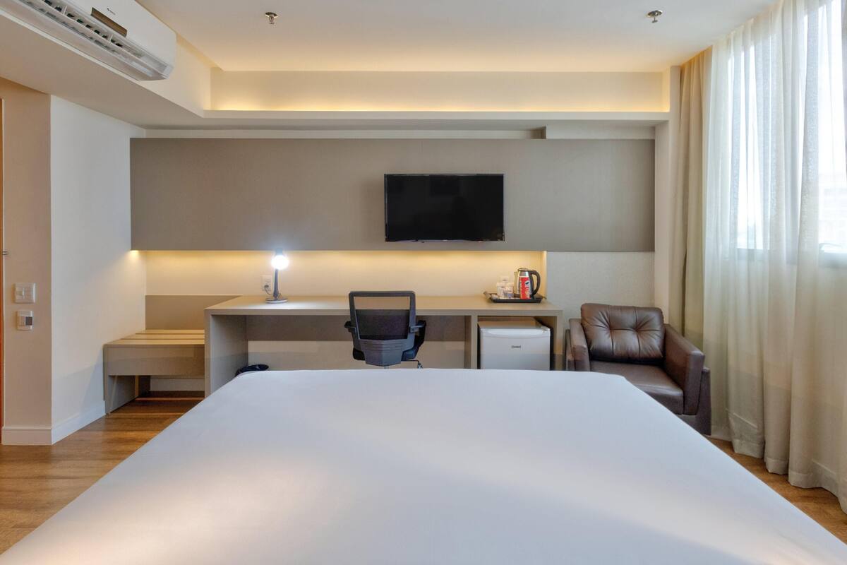 Habitación de hotel con una cama grande, un escritorio con silla, televisor, cafetera y sillón de cuero.