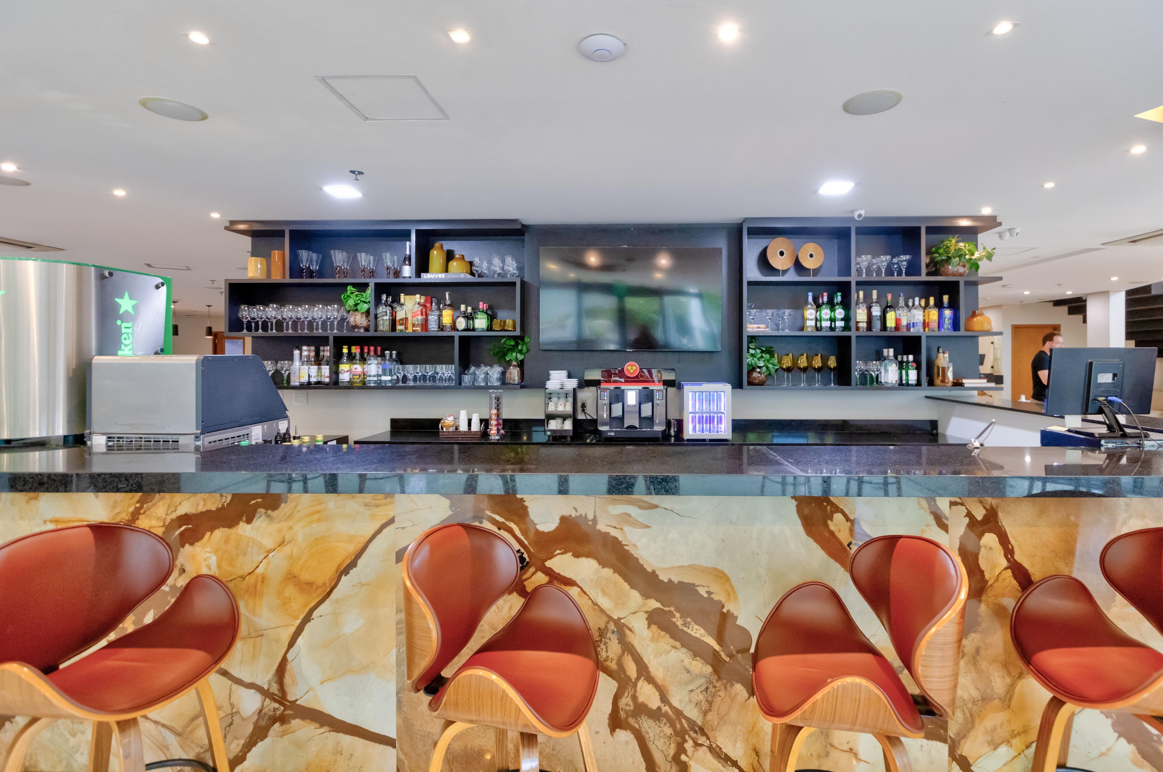 Een bar met zitplaatsen, flessen, glazen en een groot scherm.