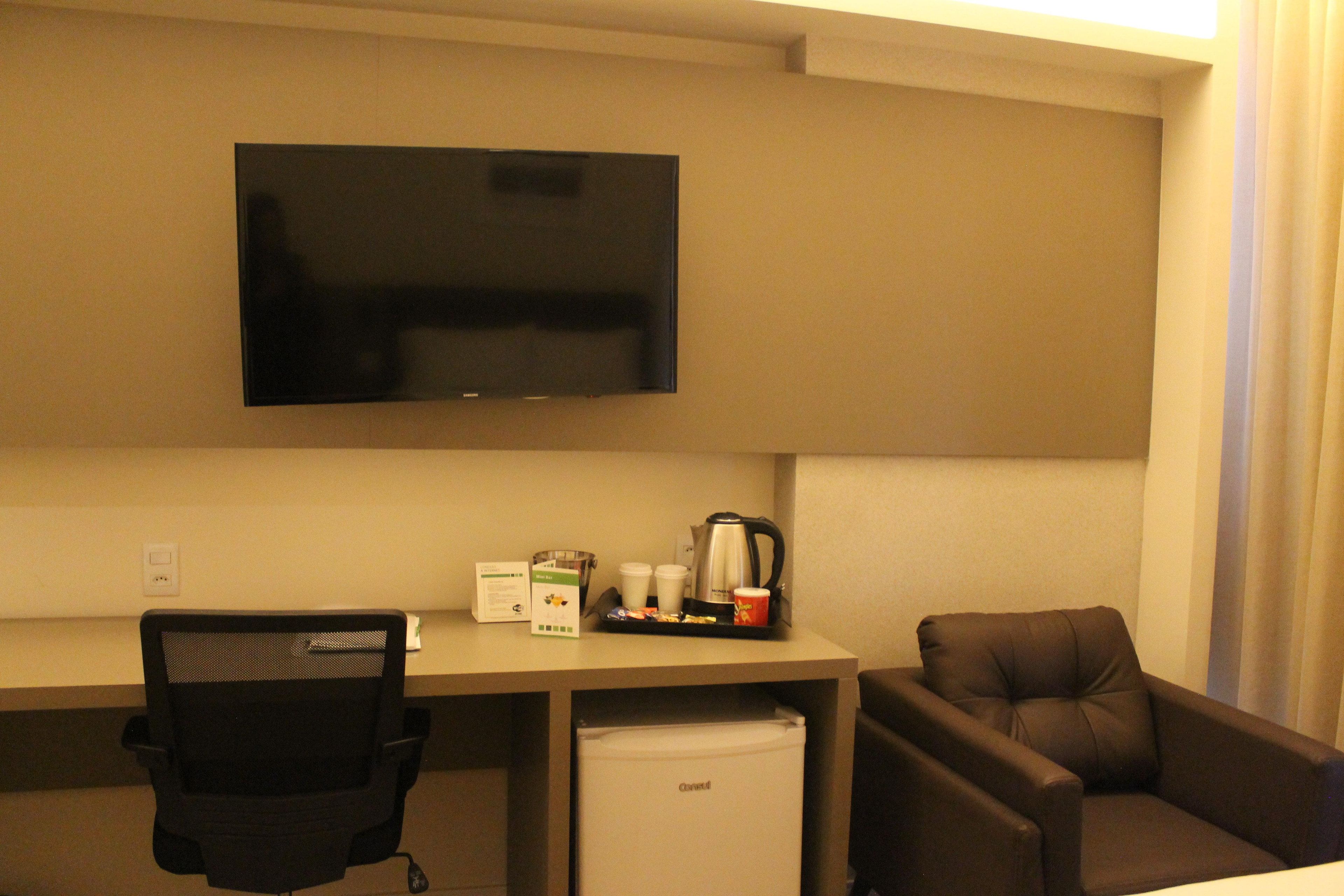 A hotel room with a flat-screen TV mounted on the wall, a desk with a coffee maker, cups, and a box of tissues. There is a black office chair and a brown armchair.