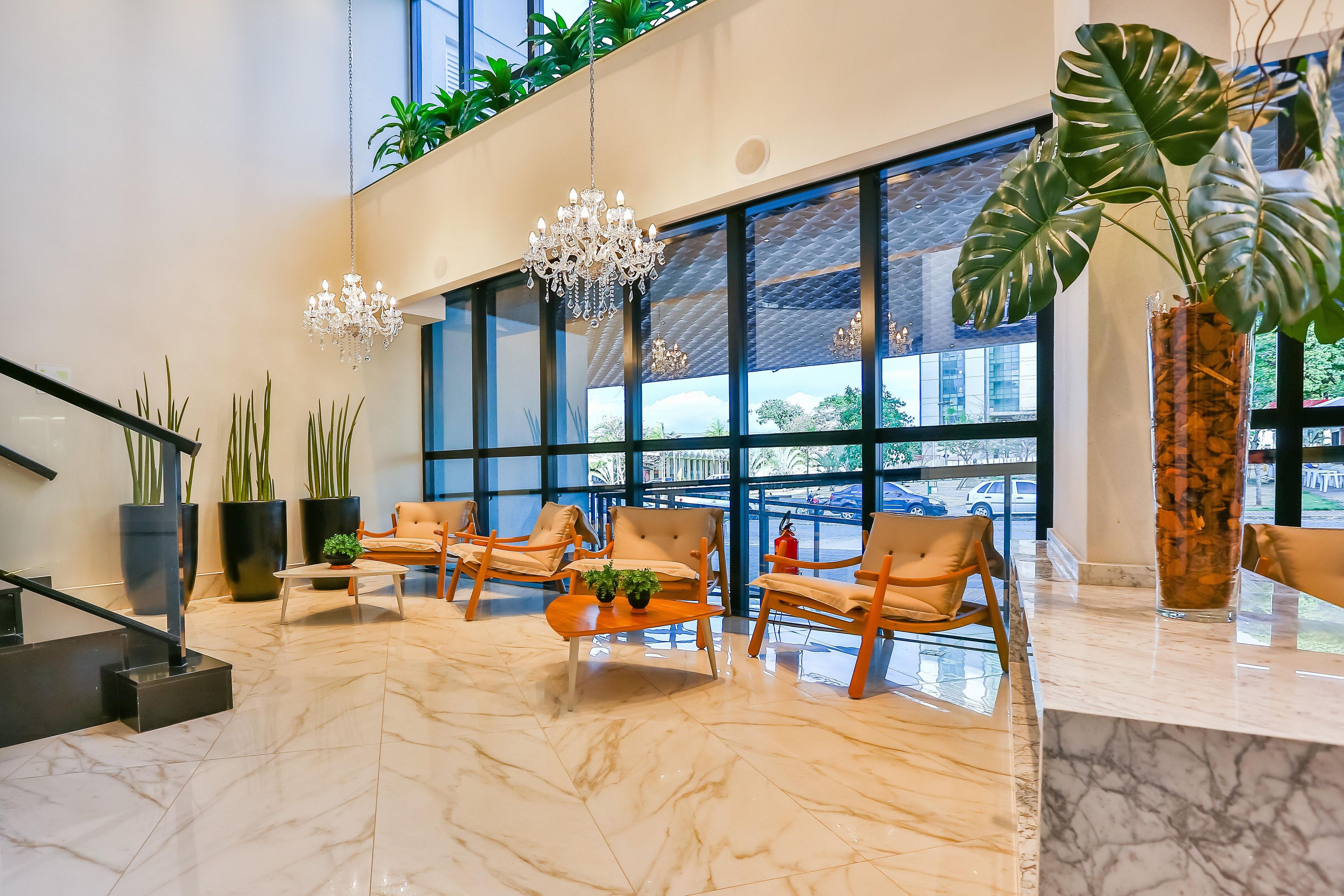 A lobby with a marble floor, wooden chairs, a chandelier, large windows, and potted plants.